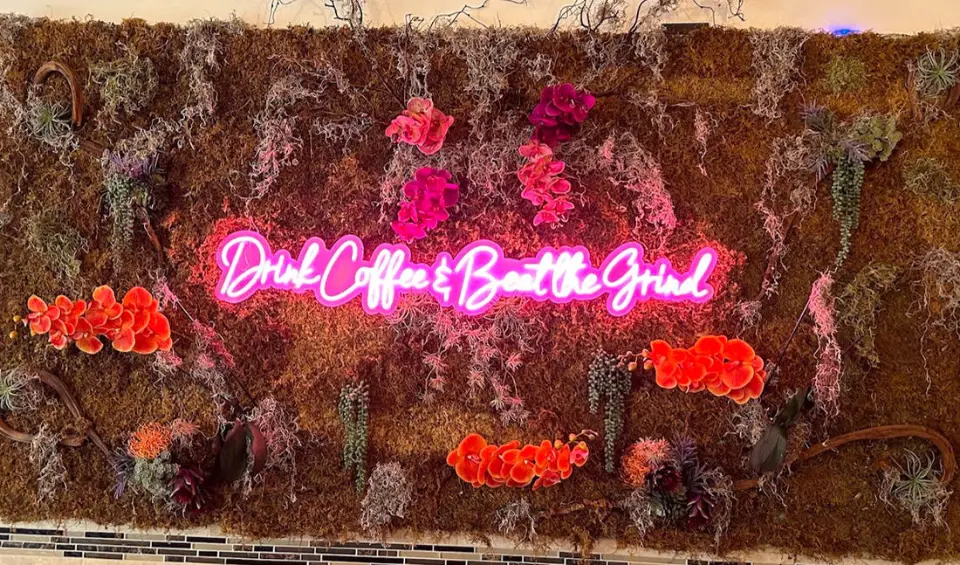 Neon sign reading Drink Coffee and Beat the Grind on a flower wall
